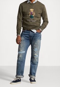 Sudadera verde oliva con un gráfico de un oso con chaqueta, combinada con jeans de mezclilla azul claro desgastados y zapatillas náuticas azul marino.