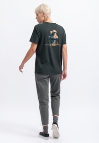 Donkergroene t-shirt met korte mouwen, voorzien van een kleurrafische graphic op de achterkant, gecombineerd met grijzige tapered broeken en zwarte instapschoenen.