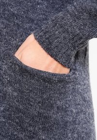 Grå ulltröja med en mjuk, texturerad yta, ribbad mudd och framficka. Tyget visar lätt variation i nyans.