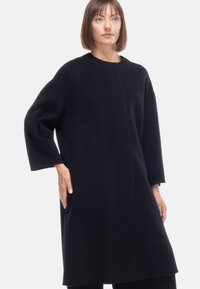 Cappotto in lana nera con un design minimalista, caratterizzato da un collo rotondo, spalle cadenti e maniche lunghe, che arriva fino a metà coscia.