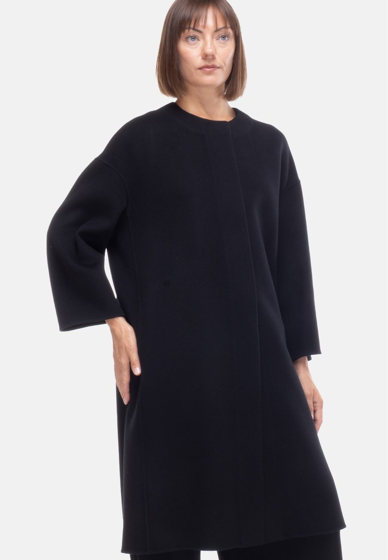 Cappotto in lana nera con un design minimalista, caratterizzato da un collo rotondo, spalle cadenti e maniche lunghe, che arriva fino a metà coscia.