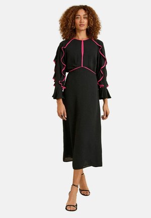 Donna che indossa un vestito midi nero con rifiniture rosa, maniche lunghe con volant e sandali neri con cinturini, in piedi contro uno sfondo neutro.