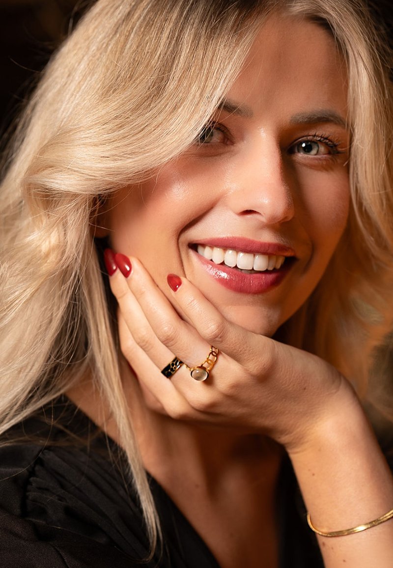Gouden ringen met een zwart accent en een steendetail. Lang blond haar, rode nagels en een zacht glimlachend gezicht. Zwarte stof op de achtergrond.