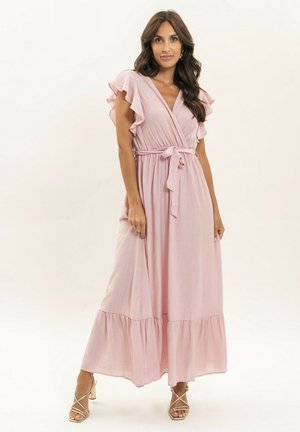 Femme debout portant une robe portefeuille rose pâle jusqu'aux chevilles, avec des manches à volants et une taille nouée, assortie à des talons dorés à brides.