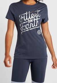 Tmavě modré bavlněné tričko s nápisem „Fittest on Earth“ ve stříbrných reflexních písmech, doplněné tmavými legínami se světle šedými akcenty.