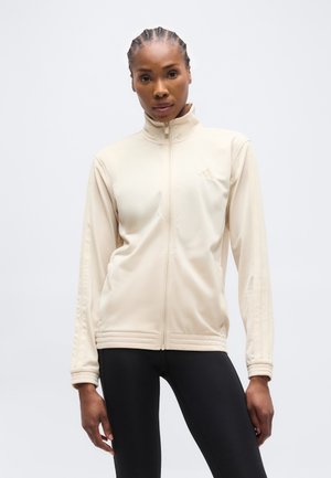 Ženska s pletenicami, oblečena v bež jakno z zadrgo in črne pajkice, stoji ob enostavni svetli podlagi.