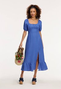 Zila linu kleita ar pūkainām piedurknēm, kvadrātveida izgriezumu, volānu apakšu un priekškopu. Modelis tur salmu somu ar ziediem. Valkāta kopā ar platformas apaviem.