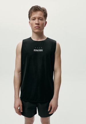 Junger Mann steht, trägt ein schwarzes ärmelloses Shirt und schwarze Shorts vor einfachem hellem Hintergrund.