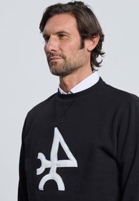 Sudadera negra con un logo abstracto blanco en el frente. Confeccionada con tela suave, cuenta con un cuello redondo y detalles de costura texturizada.