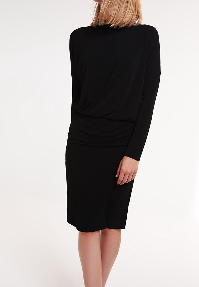 Femme portant une robe noire ajustée à manches longues avec un col montant, debout avec une main détendue le long du corps.
