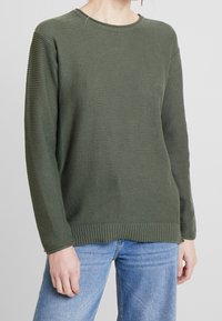 Maglione a maglia verde oliva con una trama testurizzata, scollo tondo, polsini e orlo a costine, e vestibilità rilassata. Abbinato a jeans blu.