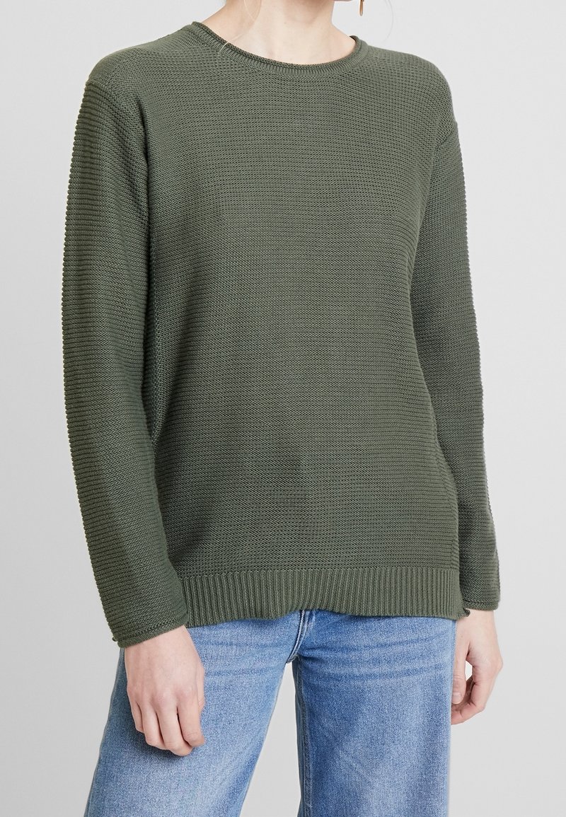 Maglione a maglia verde oliva con una trama testurizzata, scollo tondo, polsini e orlo a costine, e vestibilità rilassata. Abbinato a jeans blu.