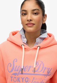 Różowa bluza z kapturem w kolorze koralowym z białymi sznurkami i nadrukowanym logo "Super Dry". Nałożona na niebieską i białą koszulę w paski z kołnierzykiem.
