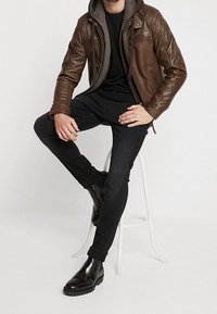 Brun læderjakke med en grå hættetrøje under, sort skjorte, sorte skinny jeans og elegante sorte ankelstøvler, siddende på en skammel.