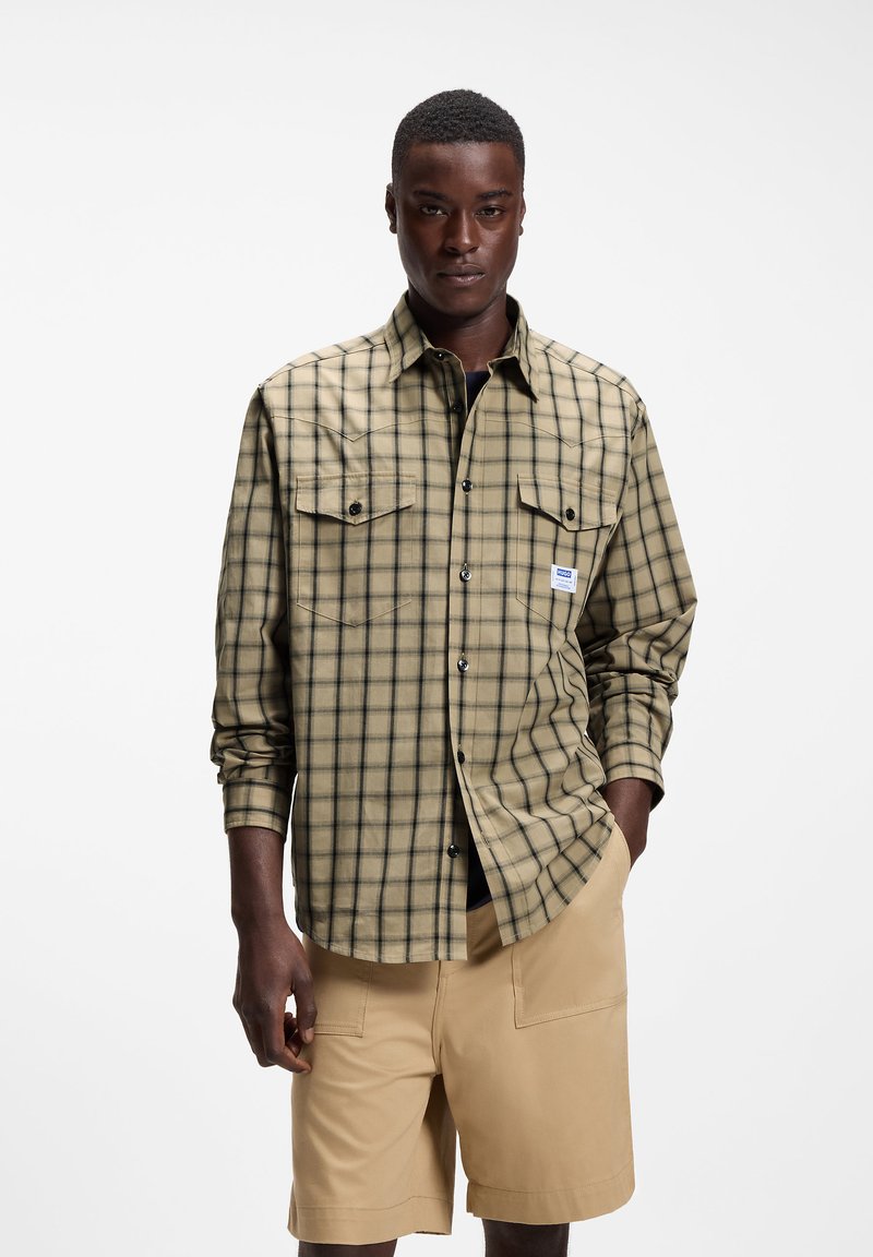 Homme portant une chemise à manches longues beige et noire à carreaux avec poches avant et un short beige, debout devant un fond blanc uni.