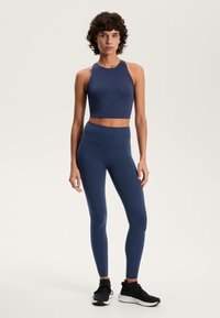 Kobieta stojąca w granatowym topie typu crop i legginsach, w czarnych tenisówkach, na jednolitym jasnym tle.