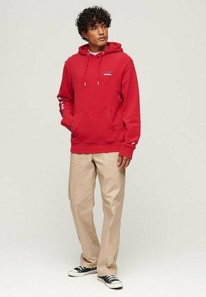 Felpa rossa con cappuccio, tasca frontale e cordino, abbinata a pantaloni beige chiaro e sneakers nere. Tessuto morbido, vestibilità rilassata.