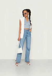 Blazer branco sem mangas sobre um top crop xadrez azul, combinado com jeans de cintura alta azul claro e sapatos de salto brancos. Acessórios incluem óculos de sol e uma bolsa branca.