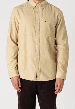 Mann trägt ein beige langärmeliges Hemd mit Knopfleiste und Brusttasche sowie dunkelbraune Hose, steht vor einem schlichten Hintergrund.