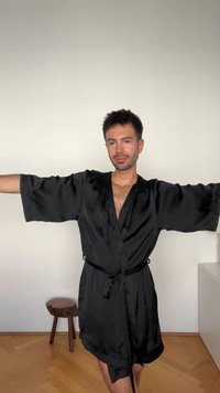 Črni satenast plašč z širokimi rokavi in zavezujočim pasom, ki ima V-izrez. Gladka tekstura z rahlim sijajem. Preprost in eleganten videz.