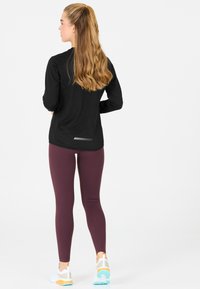 Schwarzes Langarmoberteil mit abgerundetem Saum, ausgestattet mit einem reflektierenden Streifen. Burgunderfarbene Leggings, figurbetont, kombiniert mit weißen und tealfarbenen Sportschuhen.