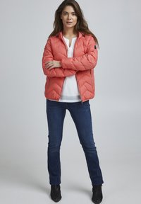Korallfarvet pufferjakke med quiltet mønster, lynlåslommer og aftagelig hætte, parret med mørke jeans og ankelstøvler.