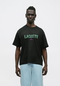 T-shirt preta de manga curta com gola redonda, com o texto "LACOSTE" bordado a verde e "Paris" a púrpura, feita de algodão, usada com calças de ganga azul claro.