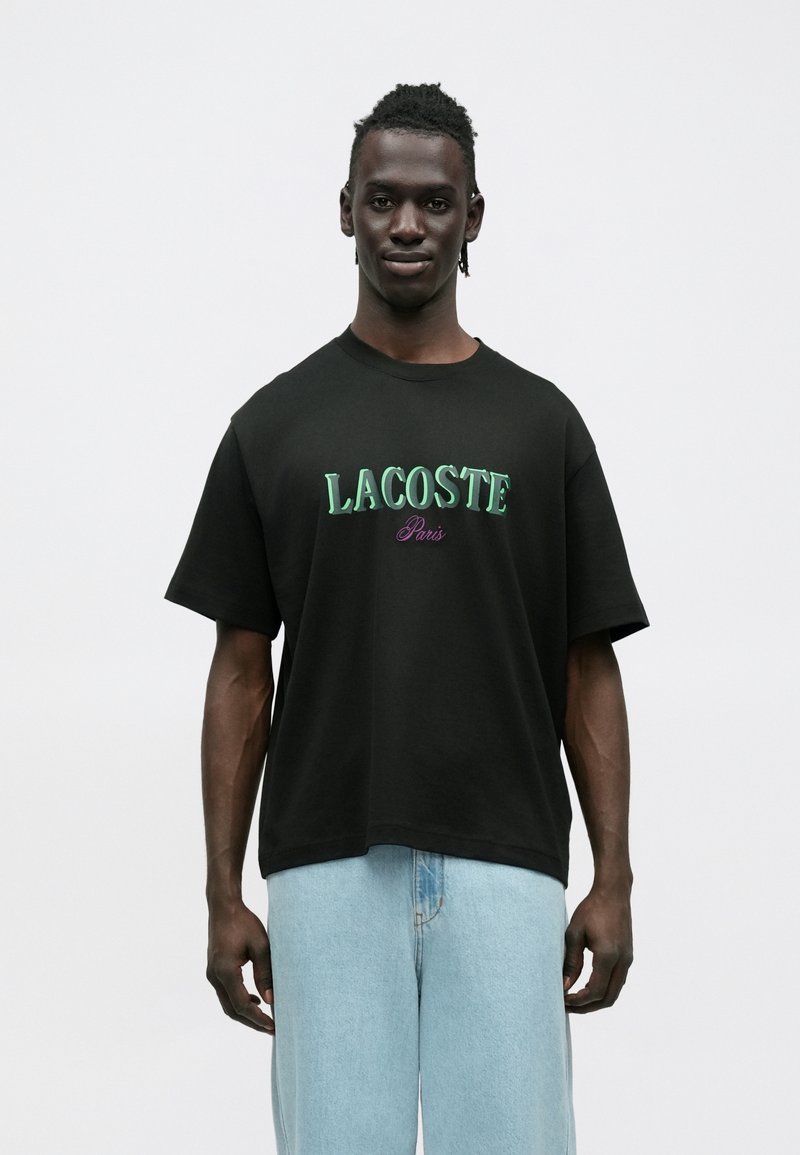 T-shirt preta de manga curta com gola redonda, com o texto "LACOSTE" bordado a verde e "Paris" a púrpura, feita de algodão, usada com calças de ganga azul claro.