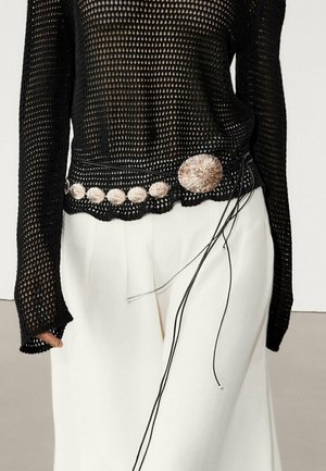 Haut à manches longues noir en maille ajourée, associé à un pantalon blanc taille haute, accessoirisé d'une ceinture ornée de pièces rondes argentées et de cordons noirs.