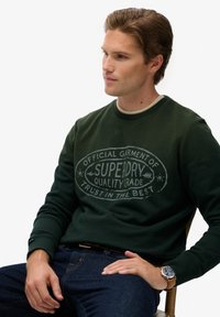 Donkergroen sweatshirt met een ronde hals en geribde manchetten, versierd met een lichtgrijze grafische print met de tekst "SUPERDRY" in het midden.
