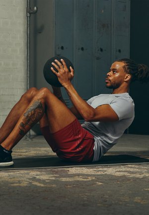 Athletischer Mann führt eine Übung auf einer Matte aus, hält einen schwarzen Medizinball, trägt ein hellgraues Shirt und rote Shorts und hat eine fitnessorientierte Haltung.