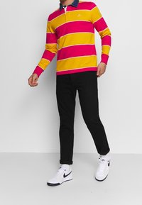 Långärmad rugby-polo med djärva rosa och gula horisontella ränder, en marinblå krage, ihop med svarta byxor och vita sneakers.