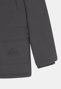 Veste imperméable noire avec une poche à rabat sur le devant, des poignets élastiques et un logo discret sur le côté inférieur gauche. Texture lisse.