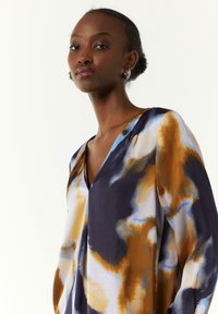 V-Ausschnitt-Bluse mit fließendem Design und kräftigen blauen, orangefarbenen und weißen abstrakten Mustern; aus leichtem, glattem Stoff gefertigt.