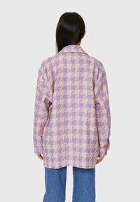 Karierte Hemdjacke in Lila und Beige, mit Kragen, langen Ärmeln und fransigem Saum. Der Stoff hat eine strukturierte Qualität.