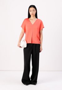 Koralā krāsas blūze ar V veida kakla izgriezumu un īsām piedurknēm, uzvilkta kopā ar melniem plašiem biksēm, turēta ar mazu baltu roku somu. Gluda auduma tekstūra.