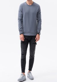 Uomo che indossa una felpa grigia a maniche lunghe, jeans neri strappati con macchie di vernice, sneakers grigie e un orologio al polso, in piedi di fronte a uno sfondo bianco.