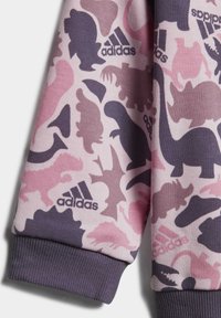 Sweatshirt violet et rose avec un motif de dinosaure, arborant un logo Adidas, des poignets côtelés et une surface en tissu texturé.