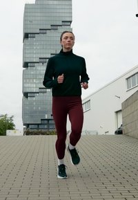 Naine jookseb ülesmäge sillutatud teel, taustal kaasaegne astmelise klaasiga hoone ja tööstuslikud struktuurid.