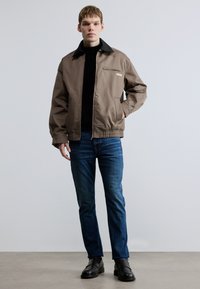 Καφέ μπουφάν bomber με μαύρο γιακά, τσέπη με φερμουάρ και ελαστική ρίγα στο τελείωμα, συνδυασμένο με σκούρο μπλε τζιν και μαύρα loafers. Λείο υφή.