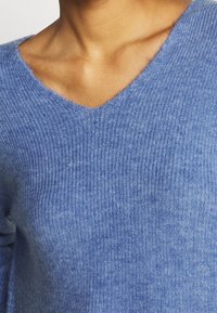 Gros plan sur une personne portant un pull en maille côtelée bleu doux avec un col en V laissant apparaître la clavicule et le haut de la poitrine.