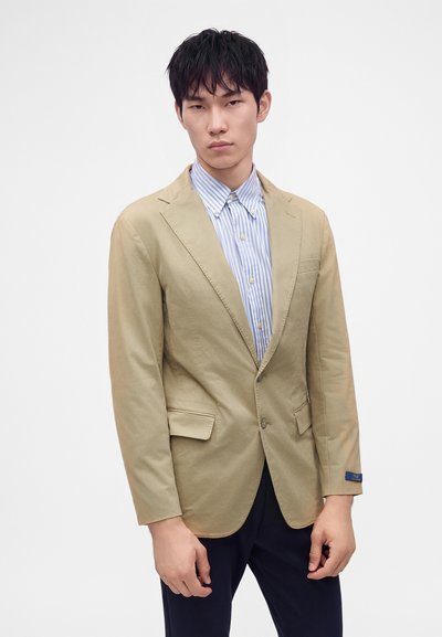 Man draagt een beige blazer over een blauw-wit gestreept overhemd met een donkere broek, staand tegen een effen witte achtergrond.