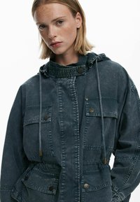 Tume denimsärk kõrge kraega, esikäetaskudega, reguleeritava kapuutsiga ja metallnuppudega; omab veidi kulunud tekstuuri.