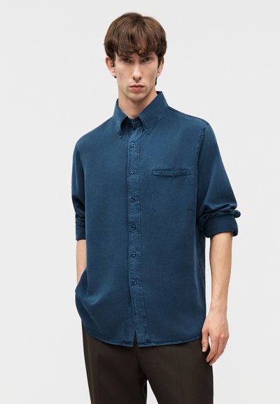 Νεαρός άνδρας με καστανά μαλλιά, που φοράει ένα μπλε πουκάμισο με κουμπιά και μανίκια που έχουν σηκωθεί, καθώς και σκούρο παντελόνι, μπροστά σε ένα απλό φωτεινό φόντο.