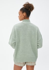 Jasnozielony, oversize'owy sweter z dzianiny z wysokim kołnierzem, prążkowanym dołem i mankietami, noszony z szortami w kratę. Miękka tekstura i luźny krój.
