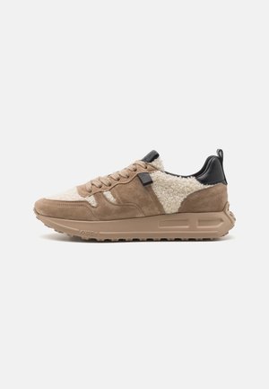 Zapatillas de ante marrón y textura crema con detalles en cuero negro, cordones redondos y una suela de goma beige gruesa.