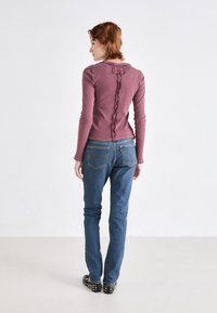 Ribbestrikket langermet topp i mauve med en bølgete bakseam og sløyfedetalj. Sammen med tettsittende blå jeans og nittede sorte sko.