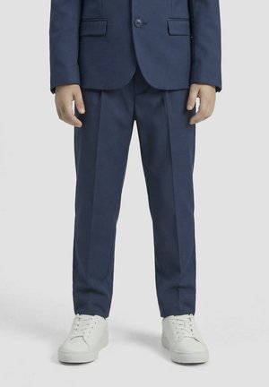 Niño que lleva pantalones y chaqueta de traje azul marino con zapatillas blancas, de pie frente a un fondo claro y liso.