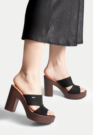 Piedi che indossano sandali neri con plateau e tacco alto, con ampie fasce, abbinati a una gonna nera al ginocchio su uno sfondo bianco.