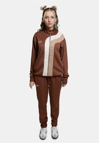 Brun treningsdrakt med beige detaljer, frontglidelås, høy krage og elastiske mansjetter. Kombinert med hvite sneakers, med grå og svarte detaljer.